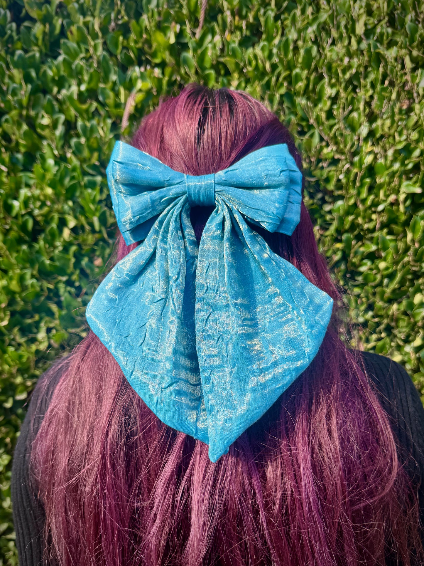 Person with red hair wearing a large light blue bow in front of a green hedge.