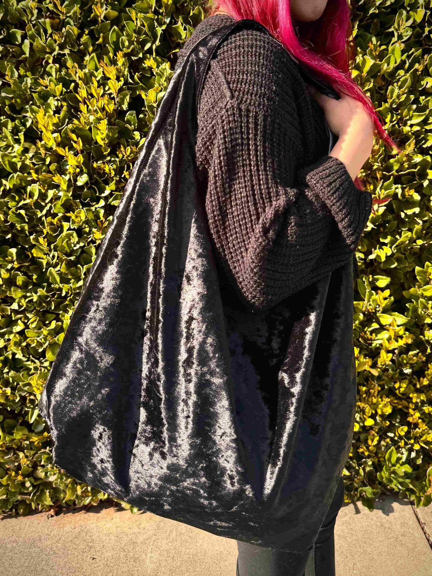Person with red hair holding a large velvet black hobo bag against a green hedge background