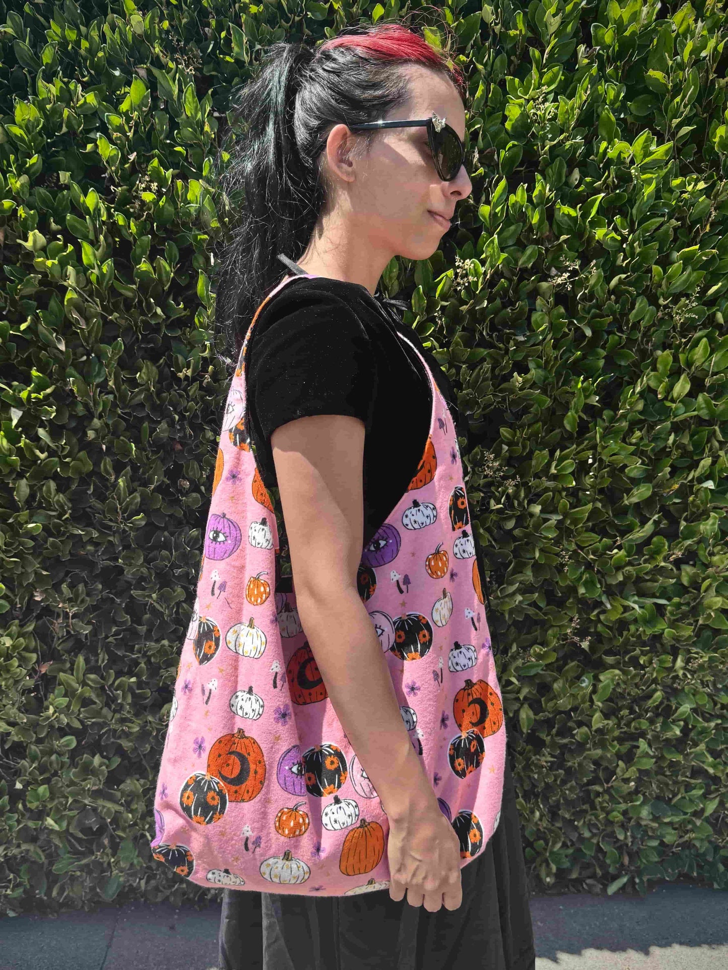 Person wearing a pink bag with Halloween-themed pumpkin design against a green hedge.
