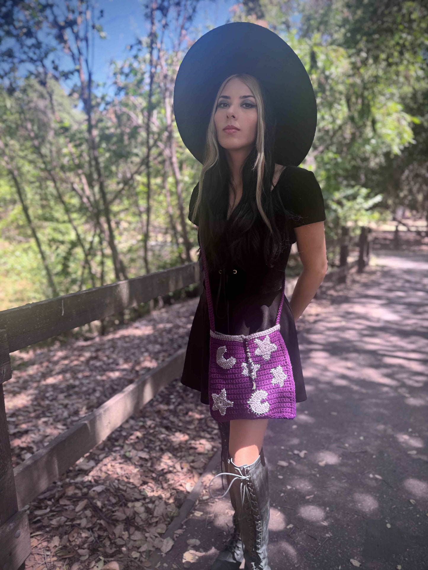 Woman in a black dress with a wide-brimmed hat and purple crochet bag with grey star and moons standing on a path with trees in the background.
