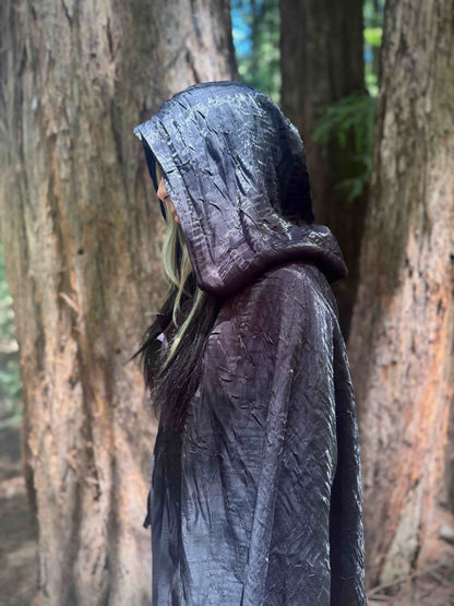 Person wearing a dark hooded cloak in a forest setting