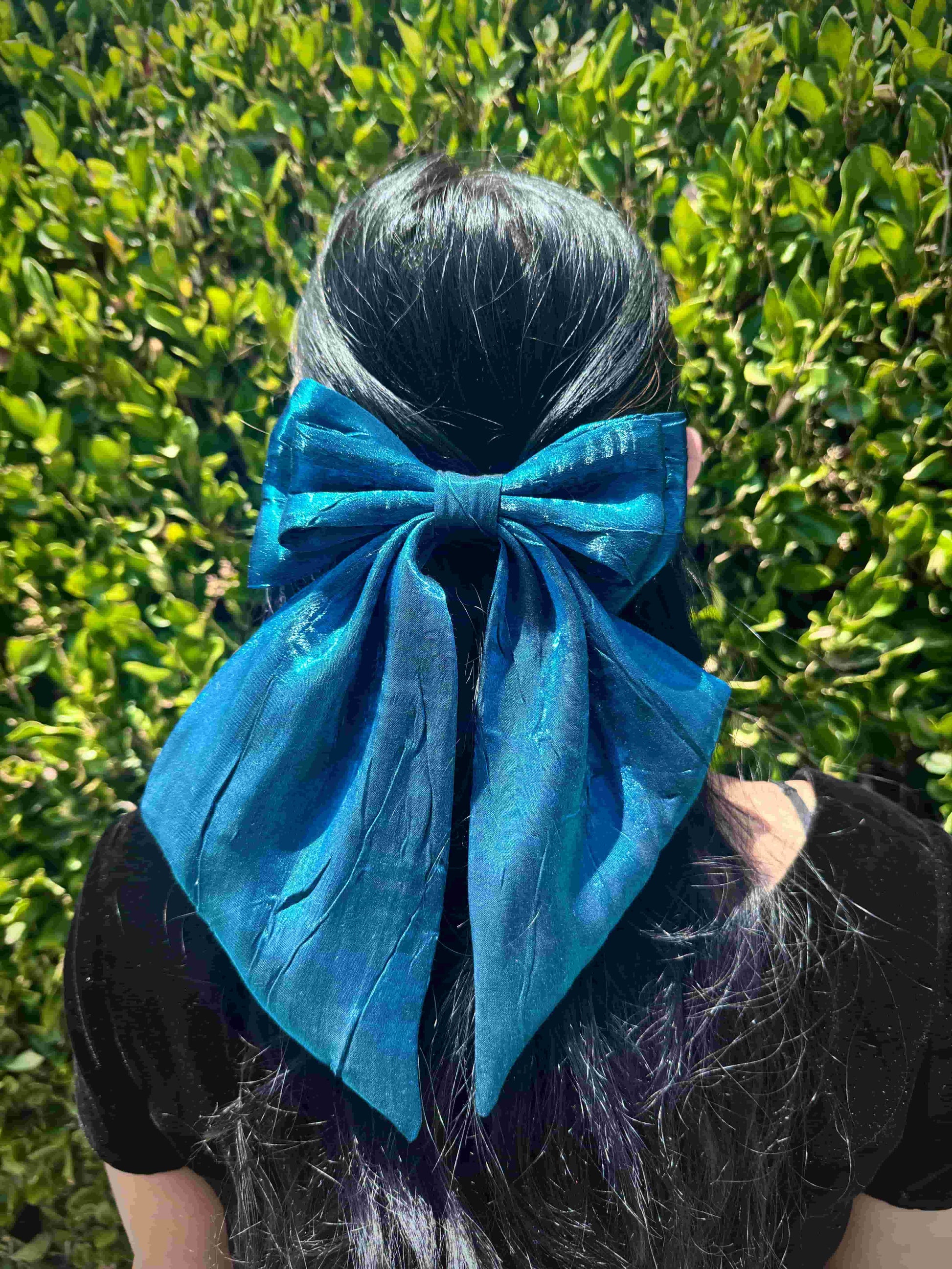 Person with a large blue bow in their hair against a green leafy background