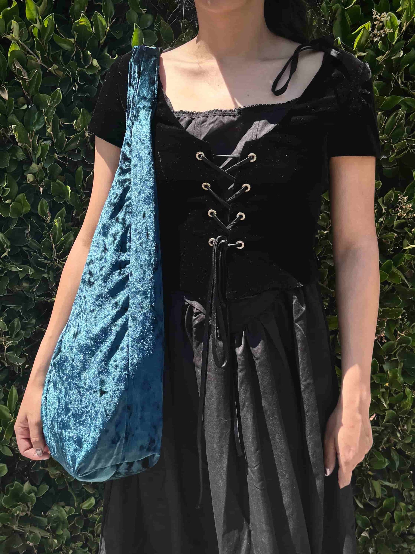 Person wearing a black outfit with a blue velvet bag over one shoulder against a green leafy background