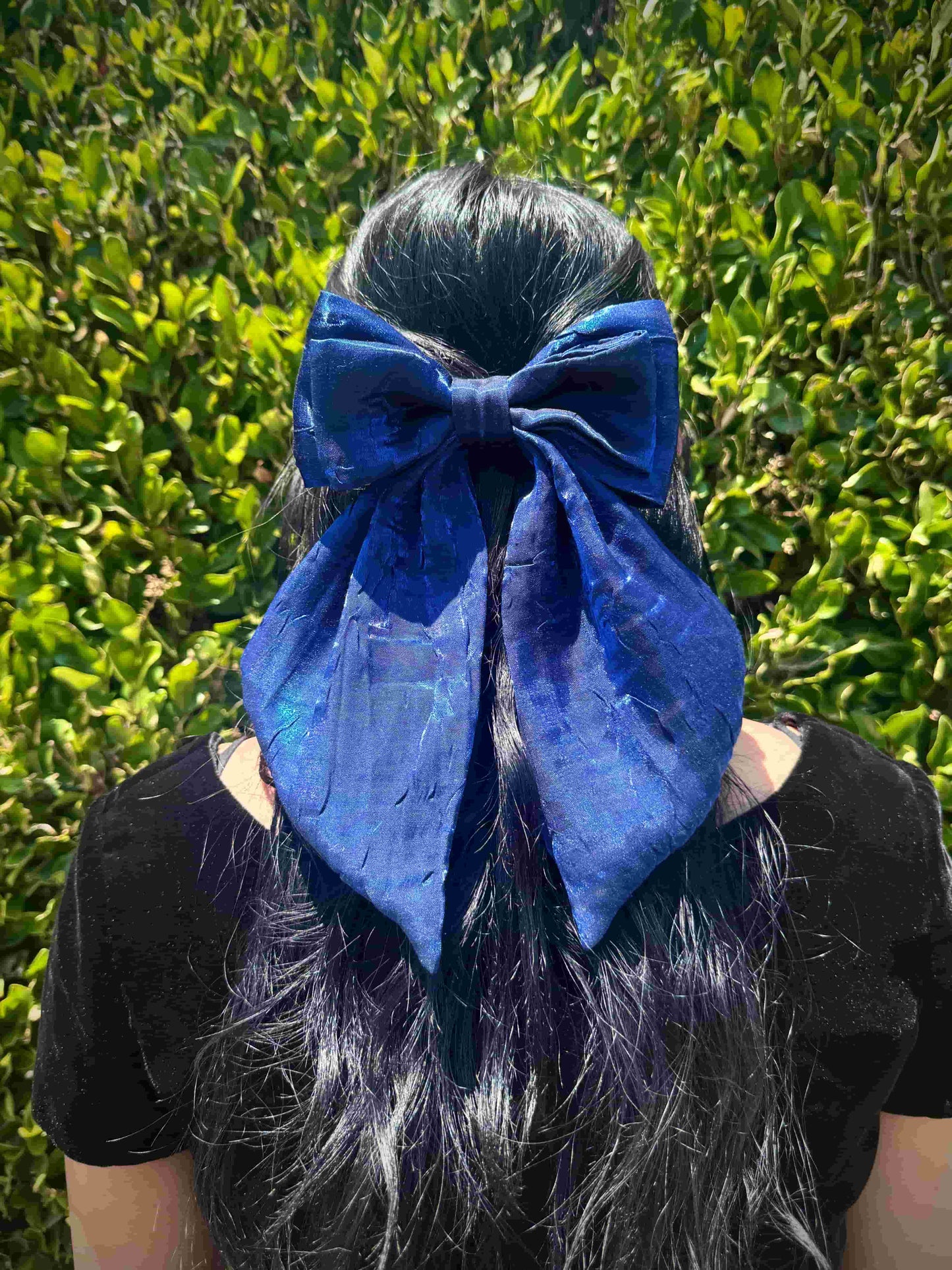 Person wearing a large blue hair bow against a green hedge background