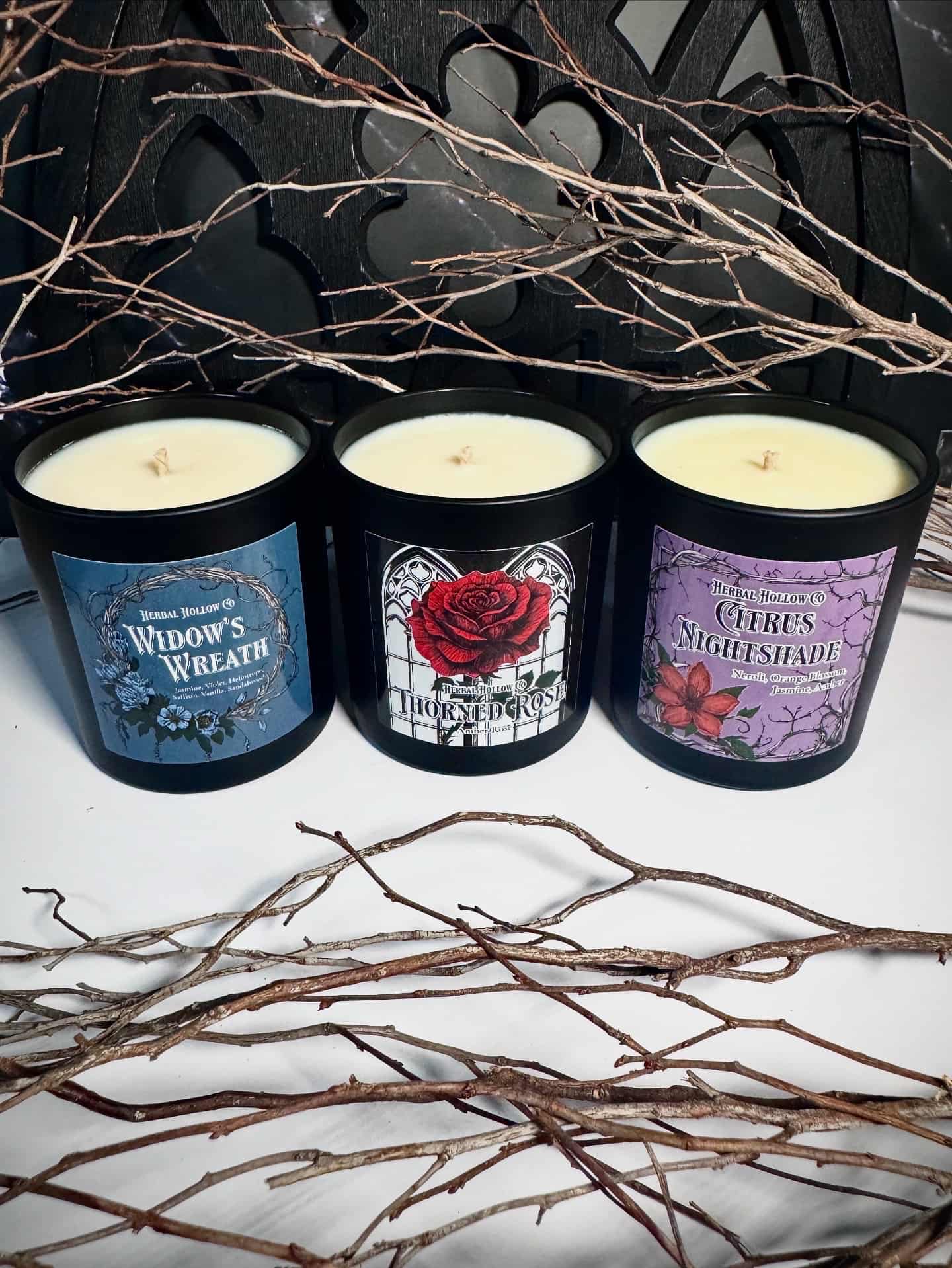 Three candles with gothic floral themed labels on a white surface with branches around