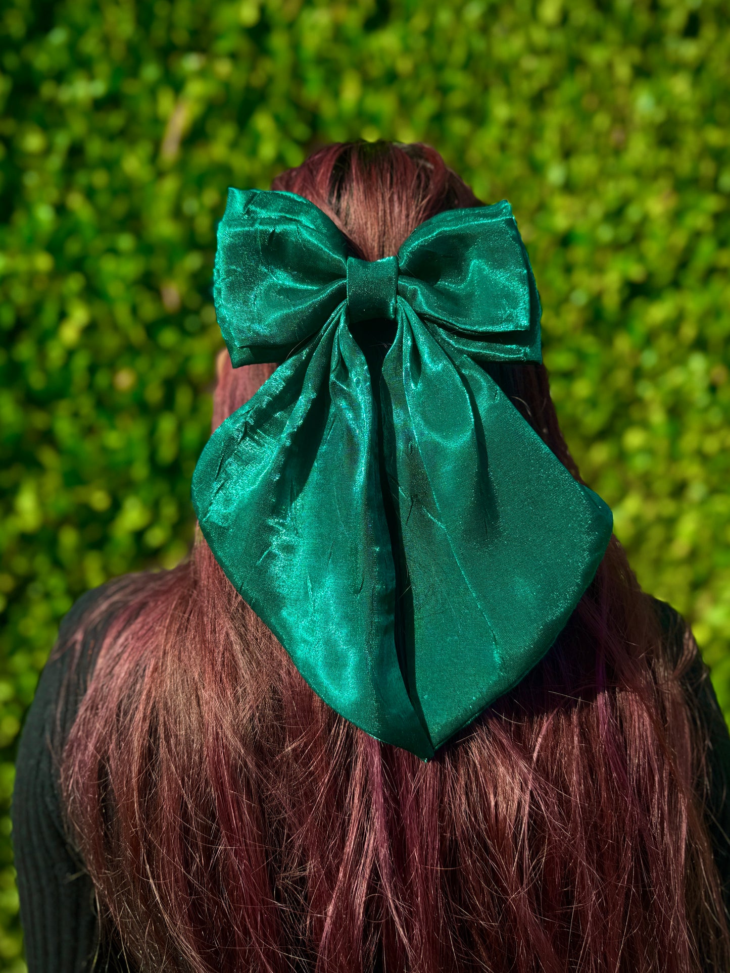 Person with red hair wearing a large dark green bow in front of a green hedge.