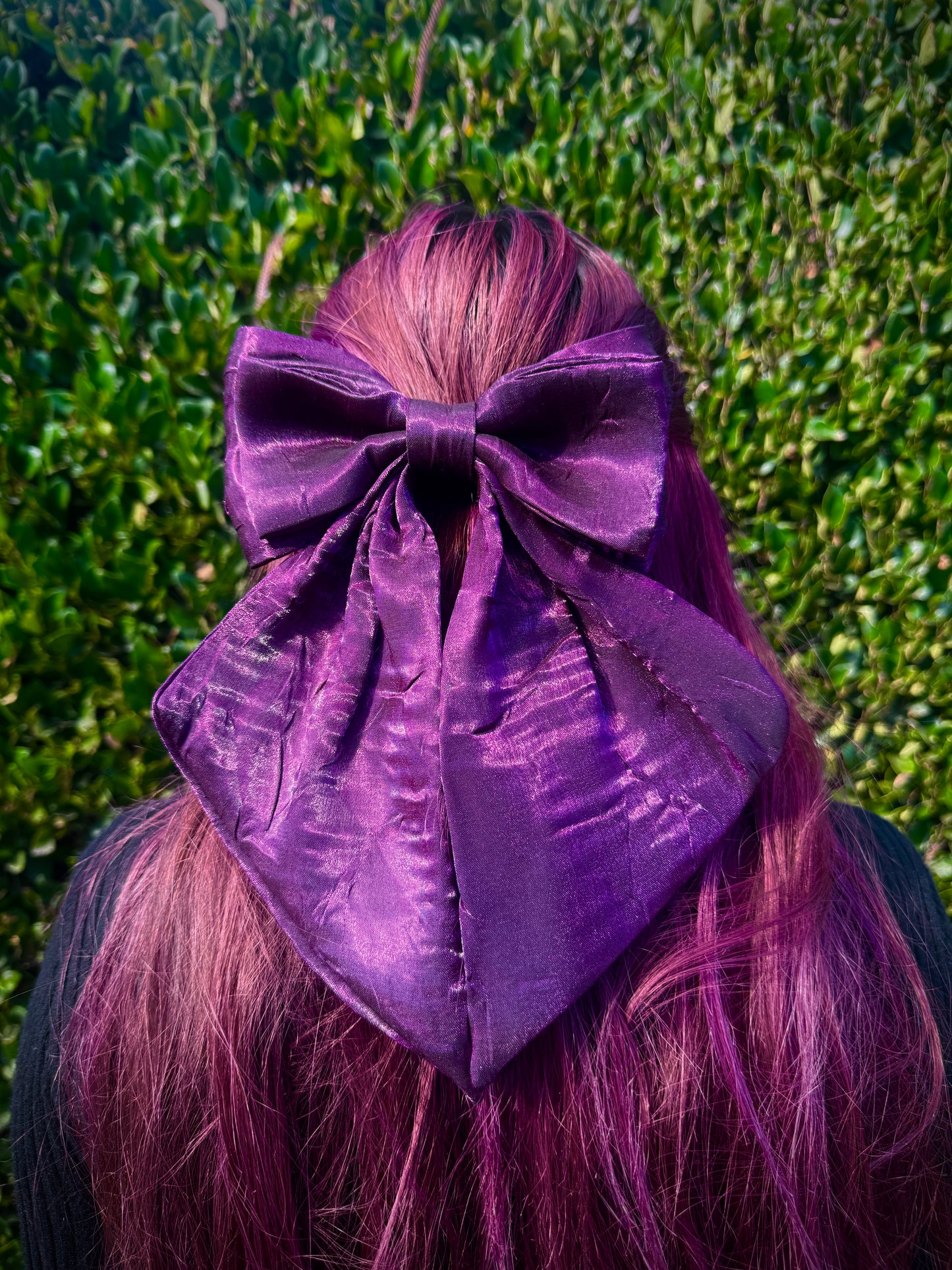 Person with red hair wearing a large purple bow in front of a green hedge.