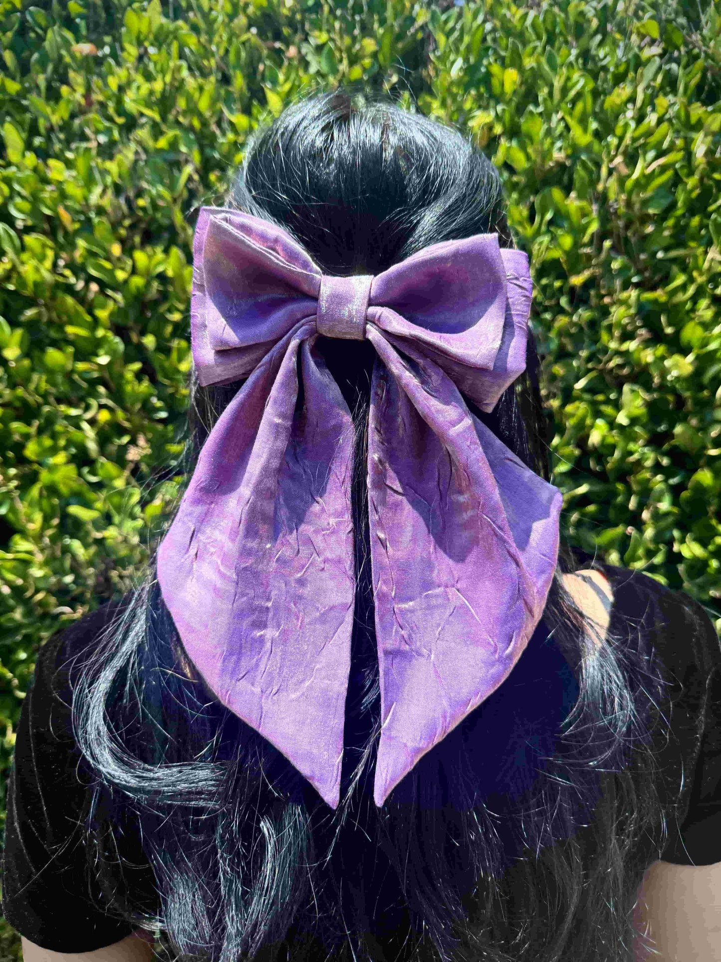Person with a large light purple hair bow against a green leafy background