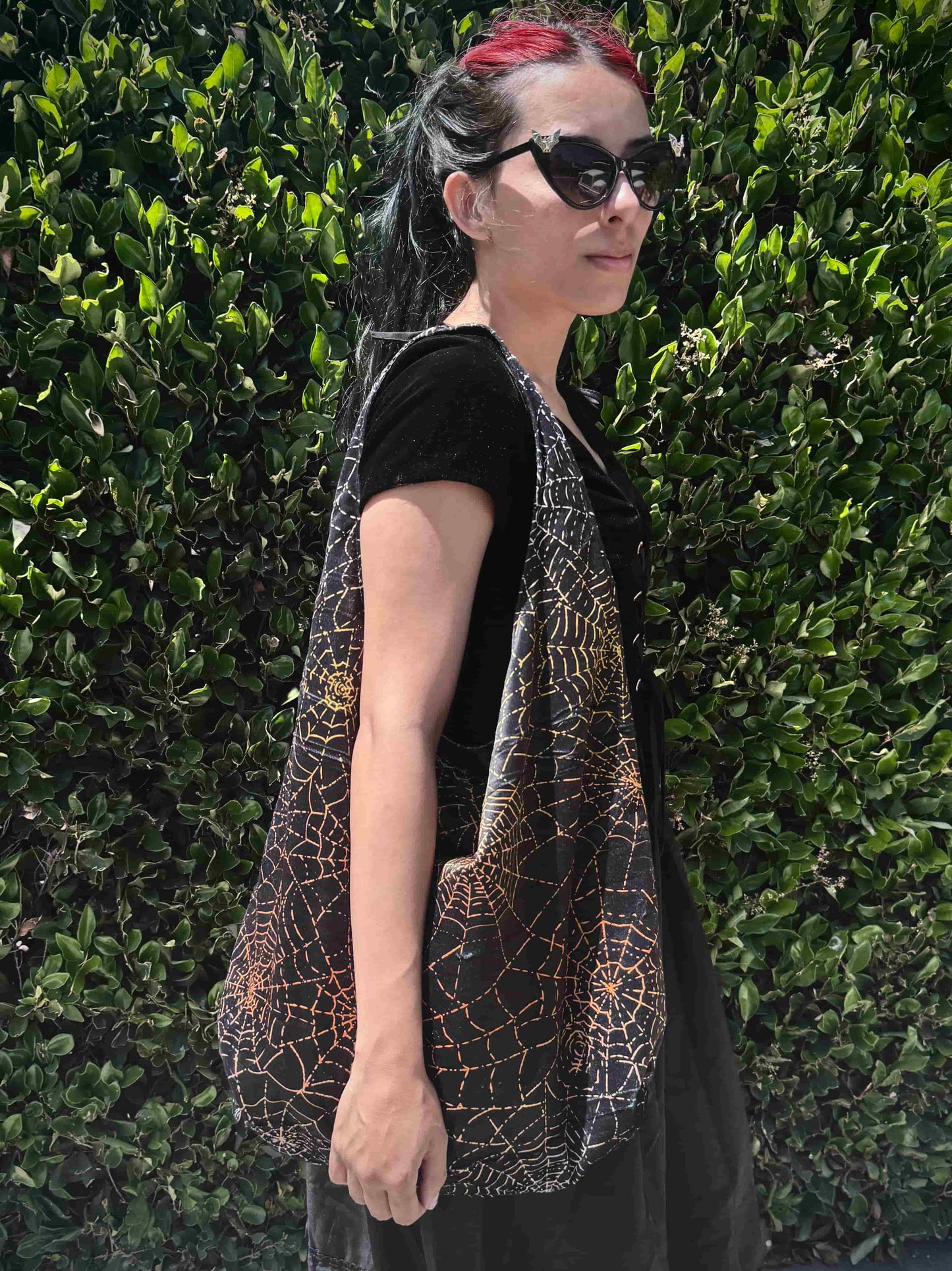 Person wearing a black and orange spiderweb patterned bag against a green hedge background