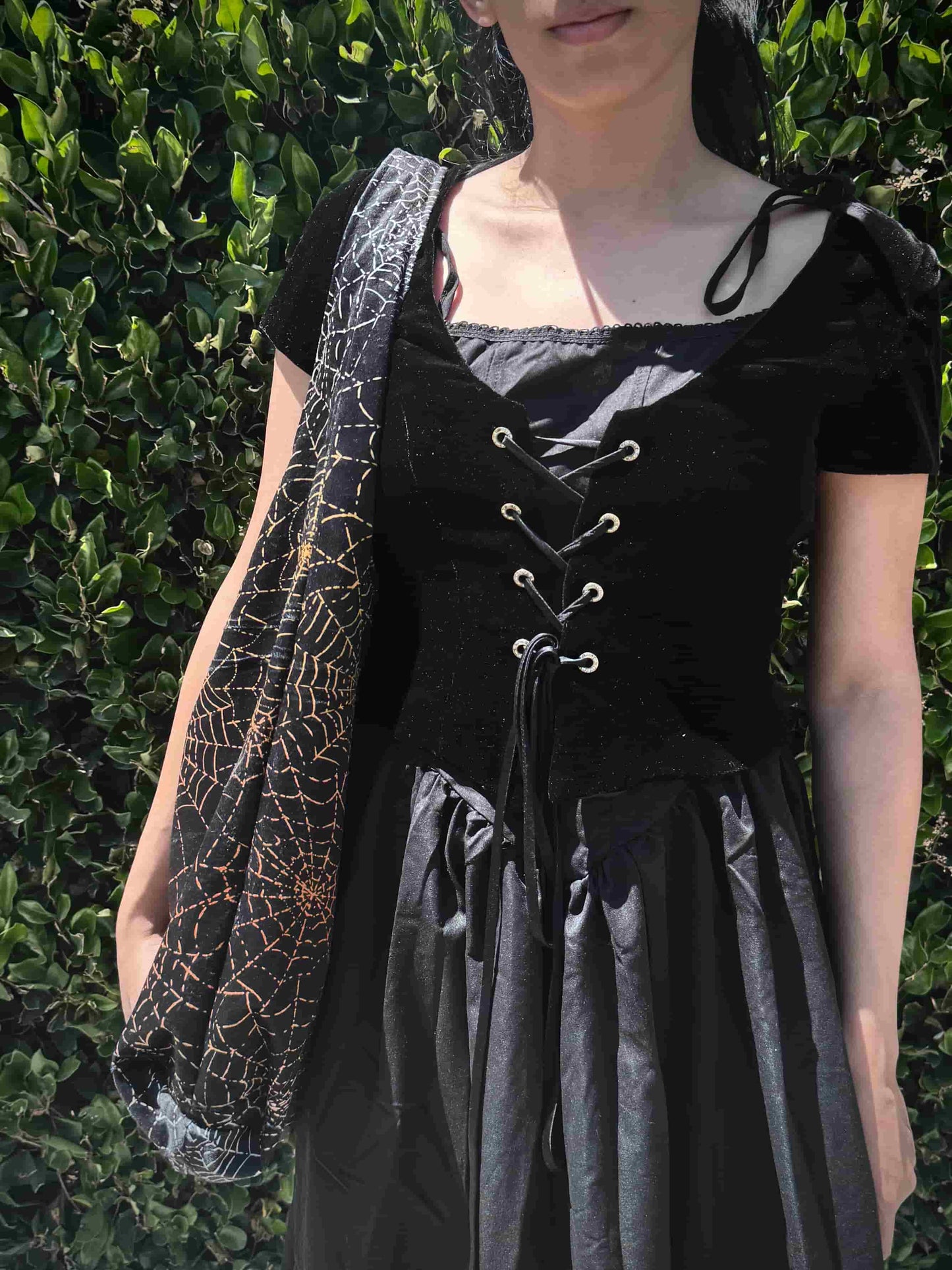 Person wearing a black and gray outfit with a spider web patterned bag against a green bush background