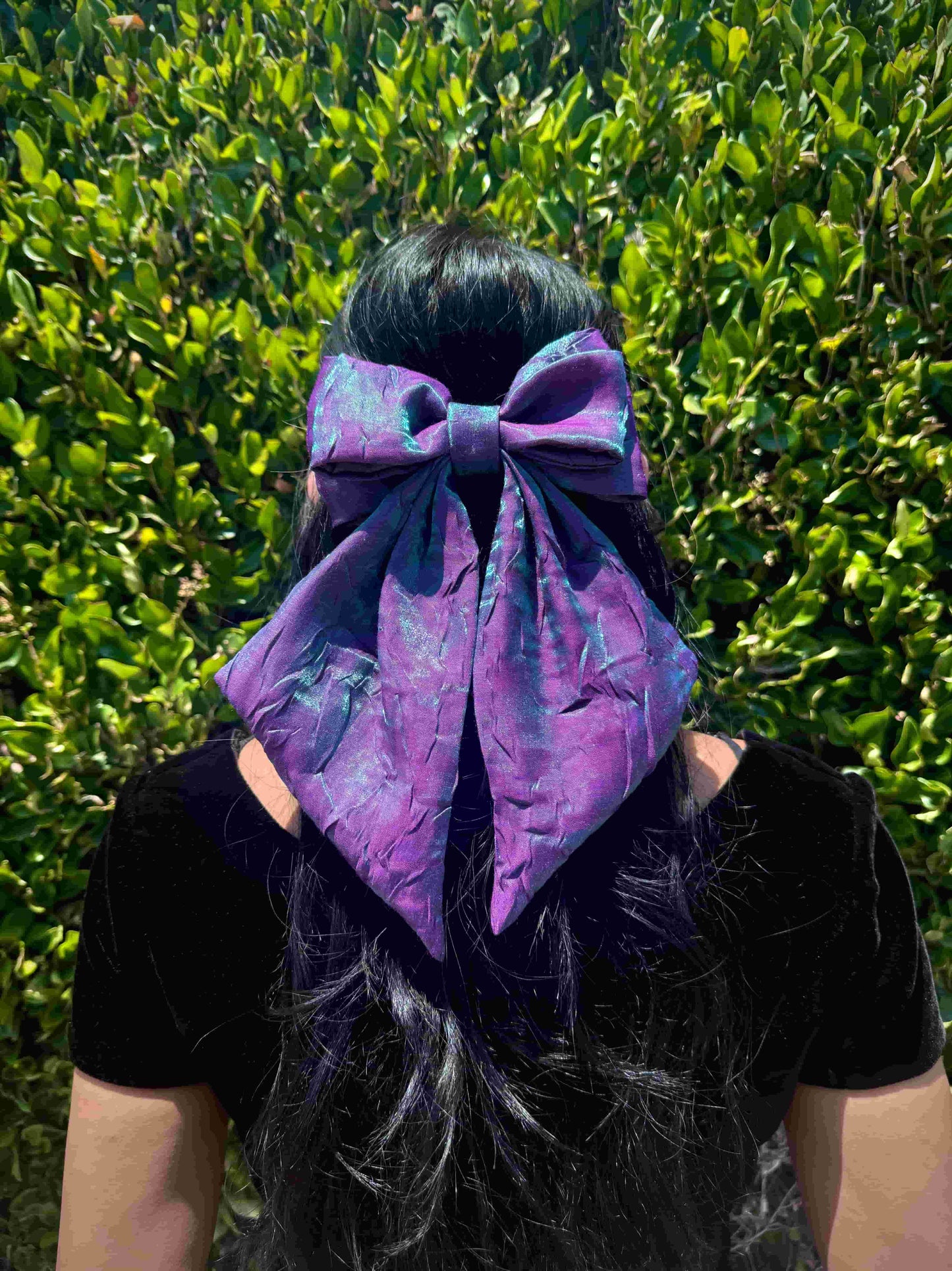 Person wearing a large two-toned blue and purple bow on their head with a green leafy background