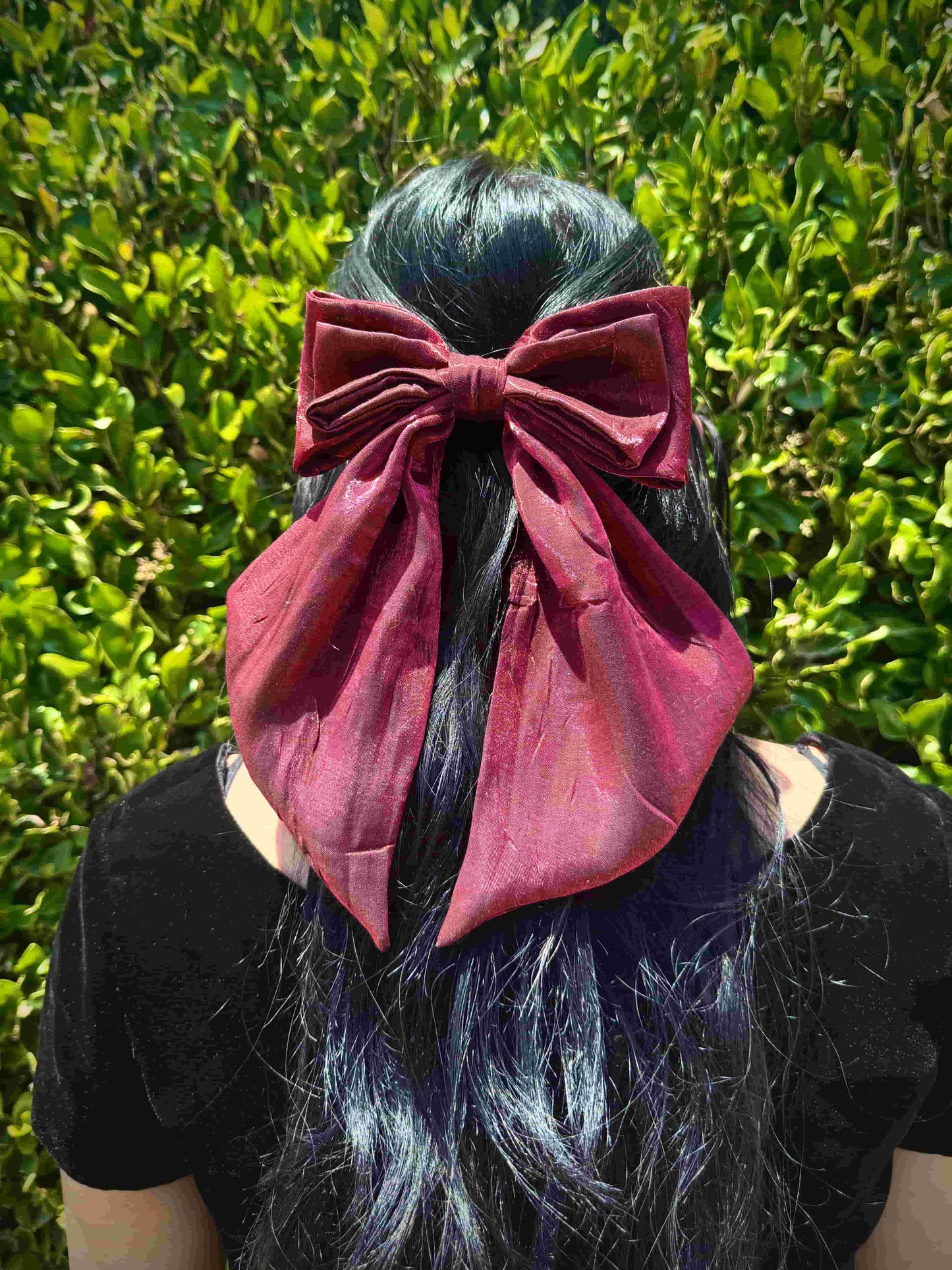 Person with a large red hair bow in their dark hair against a green leafy background