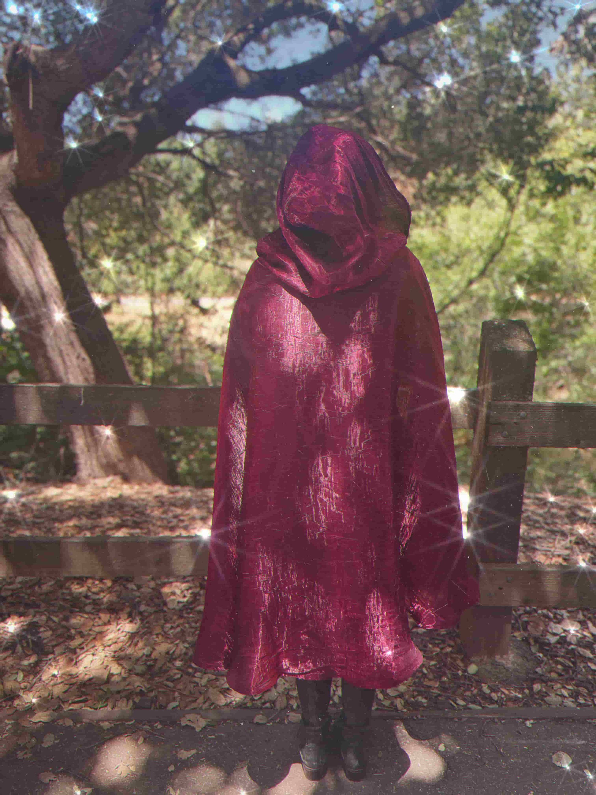 Person wearing a red hooded cape standing in a natural setting with trees and a wooden fence.