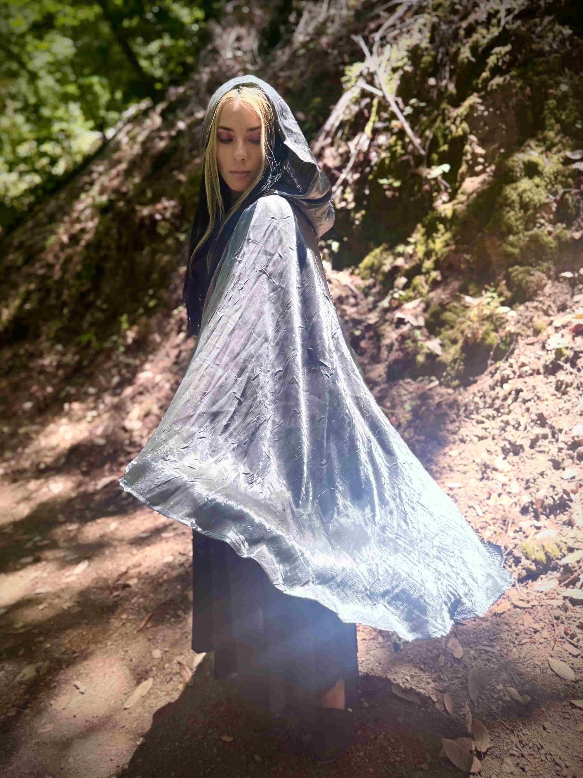 Person wearing a silver hooded cloak in a forest setting, spinning