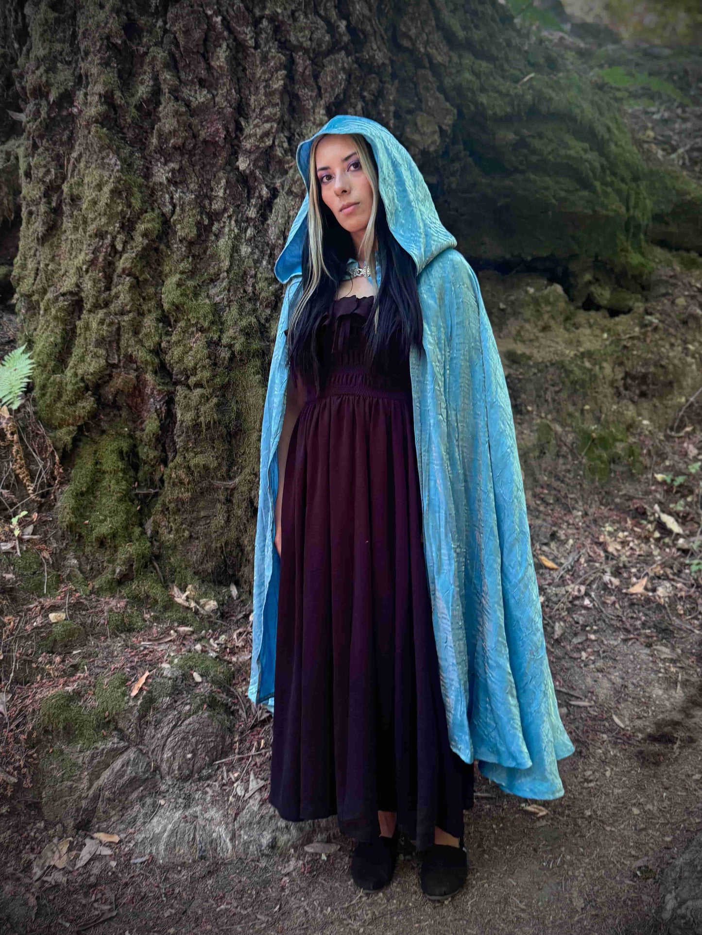 Person wearing a light blue hooded cloak and dark dress standing in a forest.