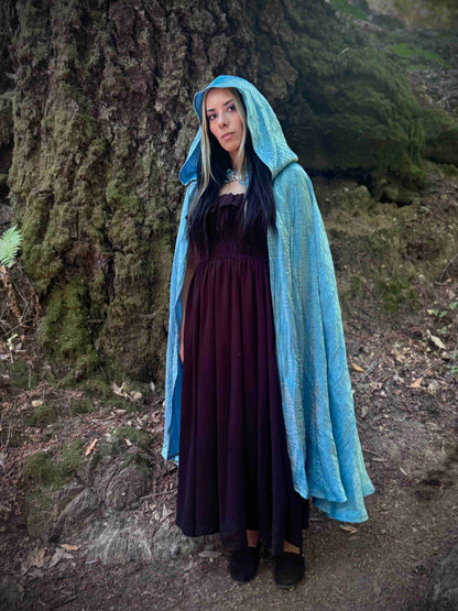 Person wearing a light blue hooded cloak and dark dress standing in a forest.