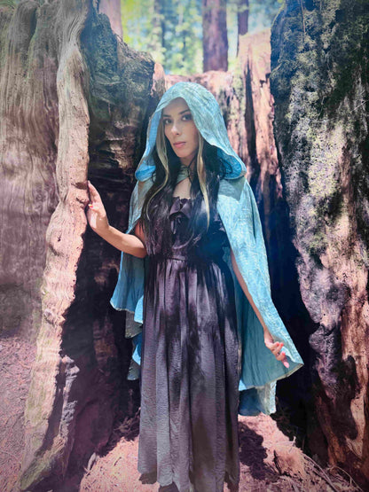Person wearing a light blue hooded short cloak and dark dress standing in a forest.