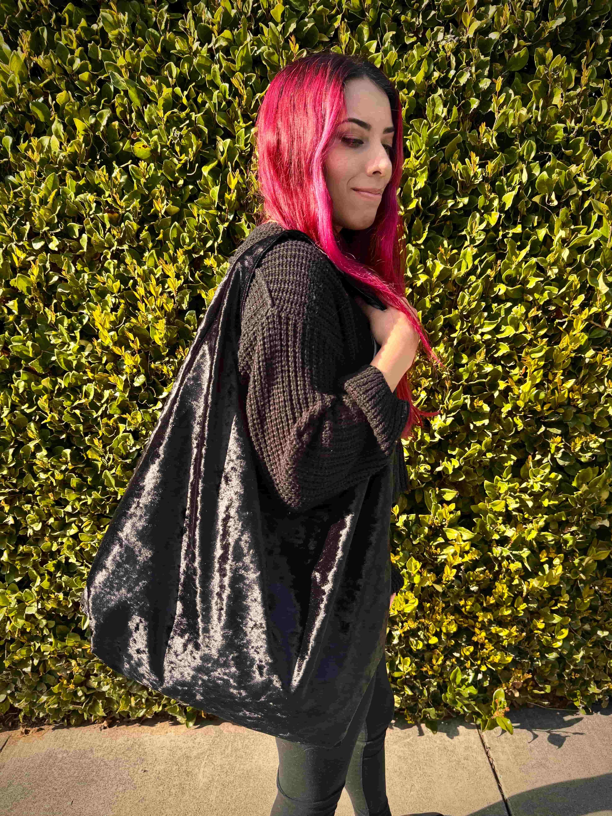 Person with red hair holding a large velvet black hobo bag against a green hedge background