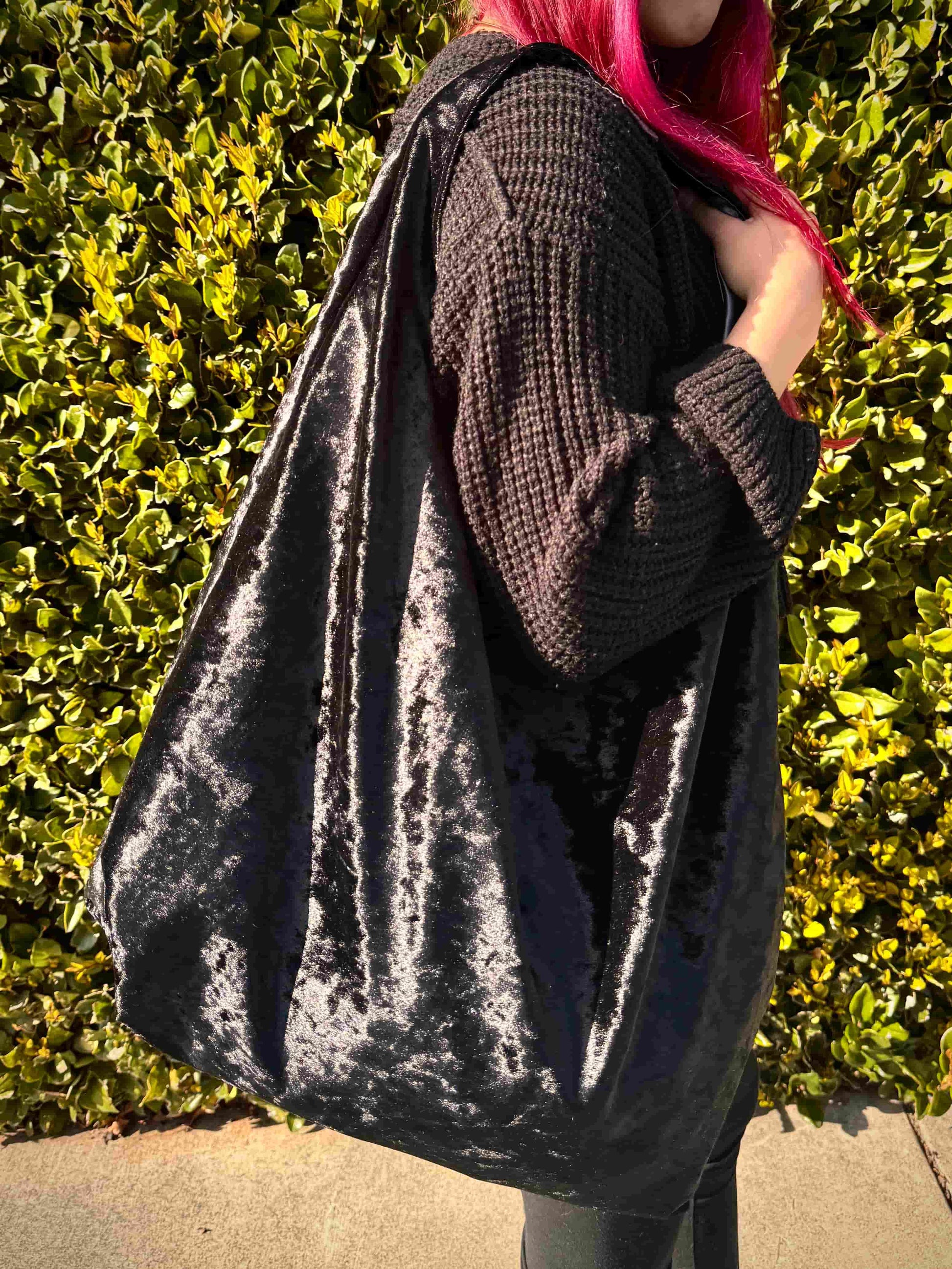 Person with red hair holding a large velvet black hobo bag against a green hedge background
