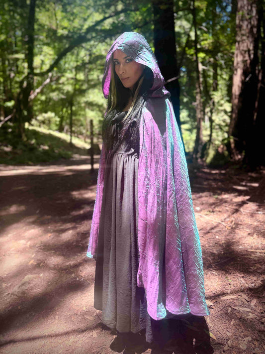Person wearing a hooded cloak with a gradient of purple and blue colors in a forest setting