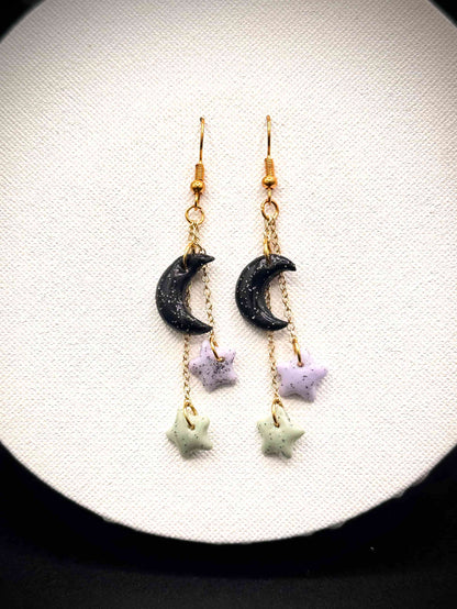 Pair of rainbow glitter black crescent moon and green and purple star earrings with gold hooks on a white background