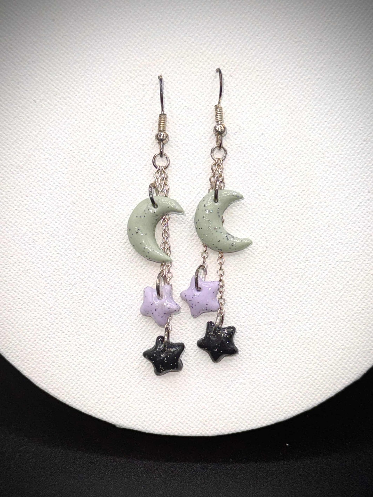 Pair of rainbow glitter green crescent moon and black and purple star earrings with silver hooks on a white background