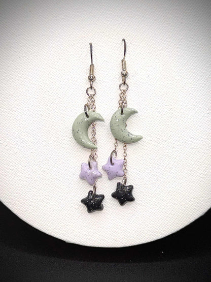 Pair of rainbow glitter green crescent moon and black and purple star earrings with silver hooks on a white background
