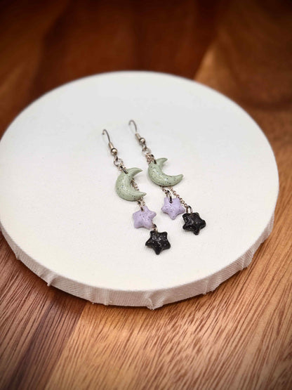 Pair of rainbow glitter green crescent moon and black and purple star earrings with gold hooks on a white and wood background