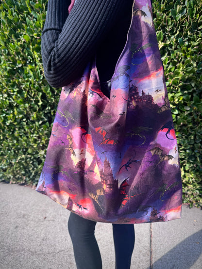 Person holding a colorful slouchy shoulder bag with a fantasy dragon and castle design against a green hedge background