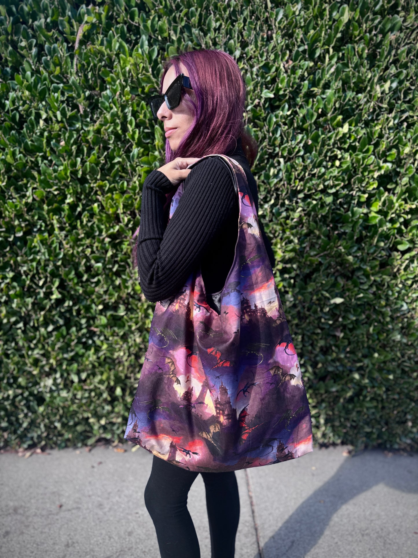 Person holding a colorful slouchy shoulder bag with a fantasy dragon and castle design against a green hedge background