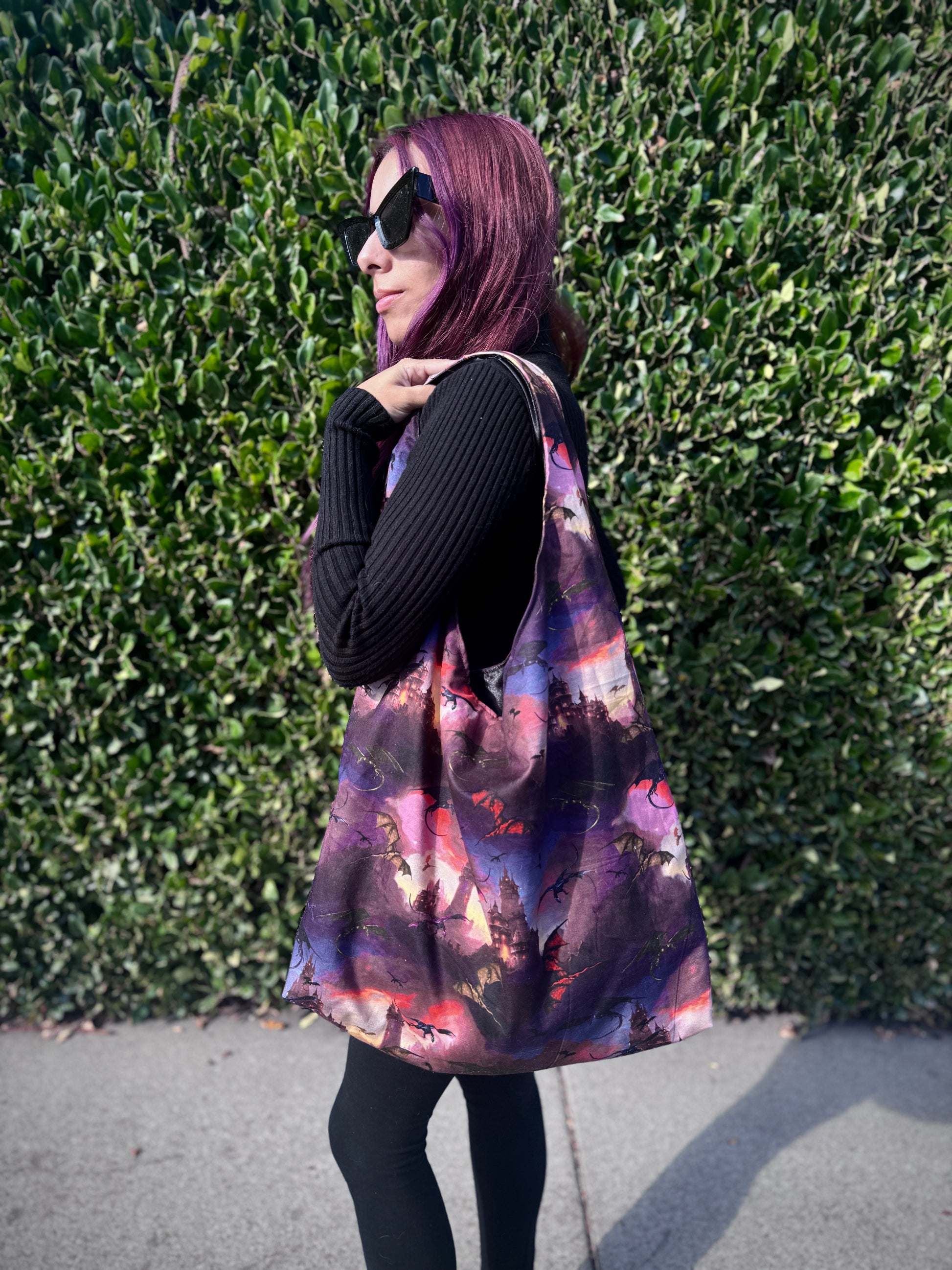 Person holding a colorful slouchy shoulder bag with a fantasy dragon and castle design against a green hedge background