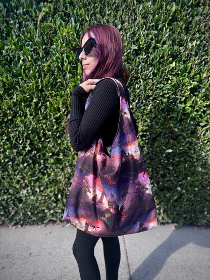 Person holding a colorful slouchy shoulder bag with a fantasy dragon and castle design against a green hedge background