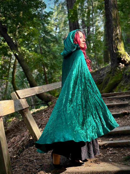 Person wearing a dark teal hooded cloak in a forest setting