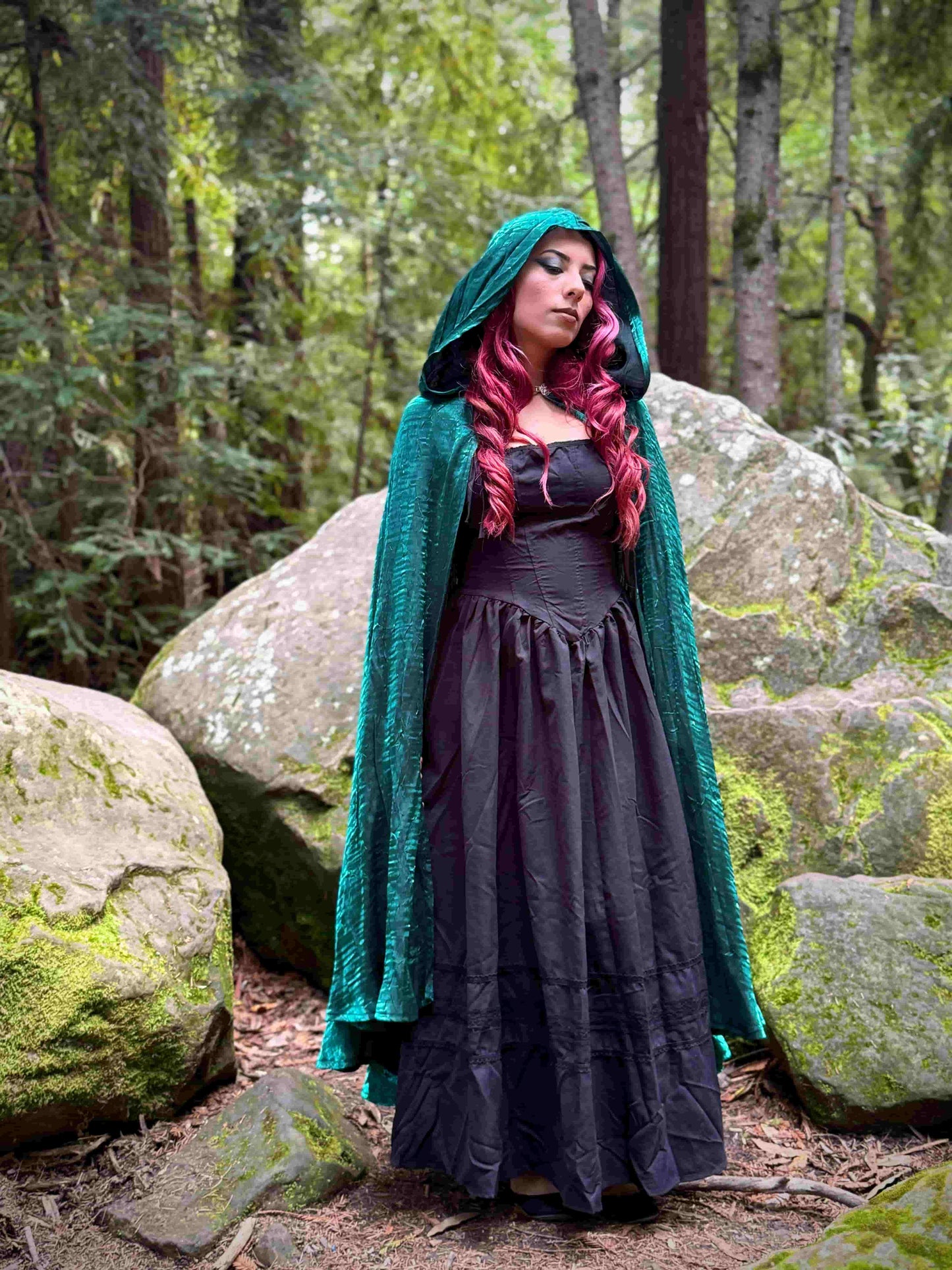 Person in a green hooded cloak and dark dress standing in a forest with large rocks and trees.