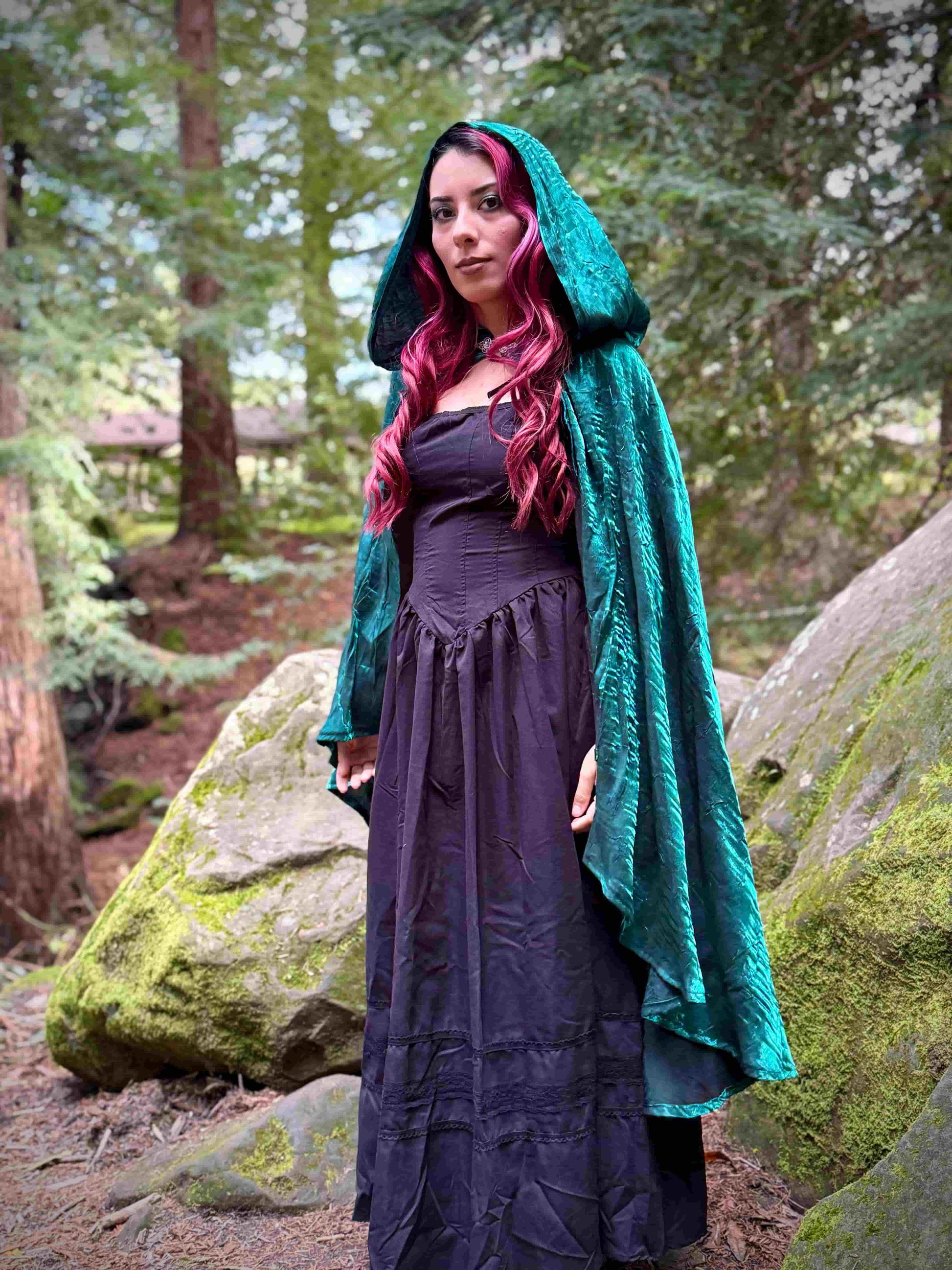 Person wearing a short dark teal hooded cloak in a forest setting