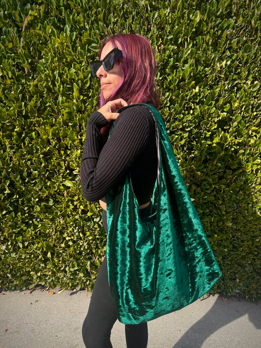 Person holding a green velvet slouchy shoulder bag against a green hedge background