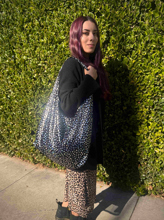 Person holding a large black and holographic stars bag against a green hedge.