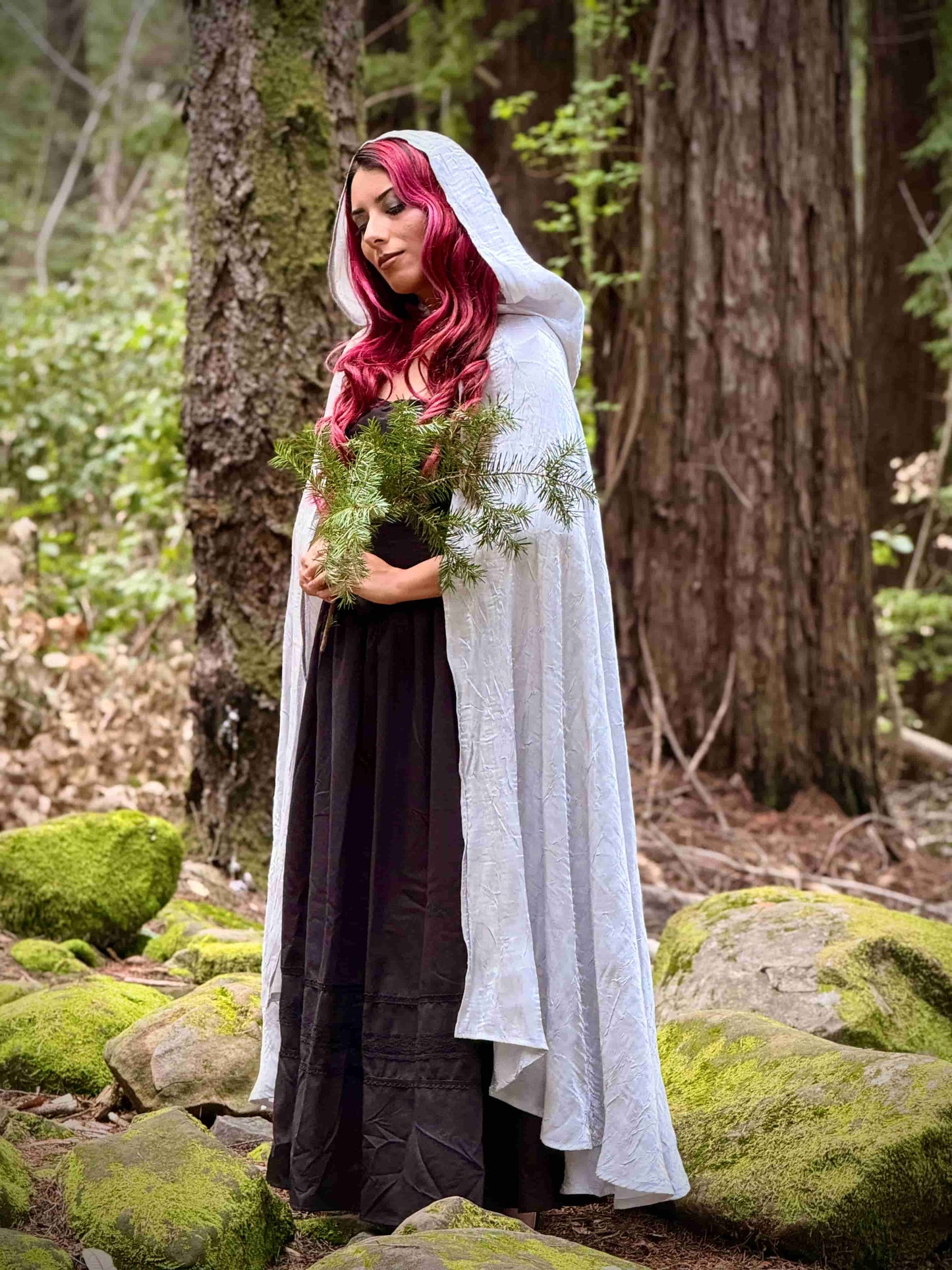 Woman in a white hooded cloak holding a plant in a forest setting