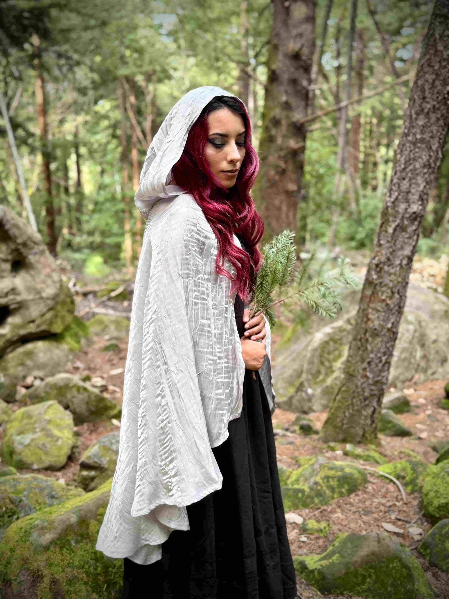 Woman in a short white hooded cloak holding a plant in a forest setting