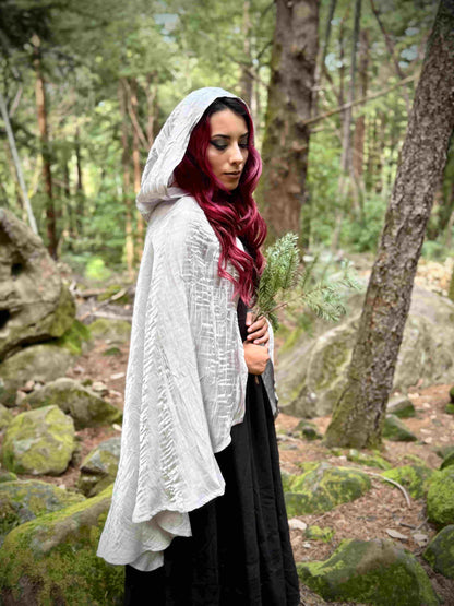 Woman in a short white hooded cloak holding a plant in a forest setting