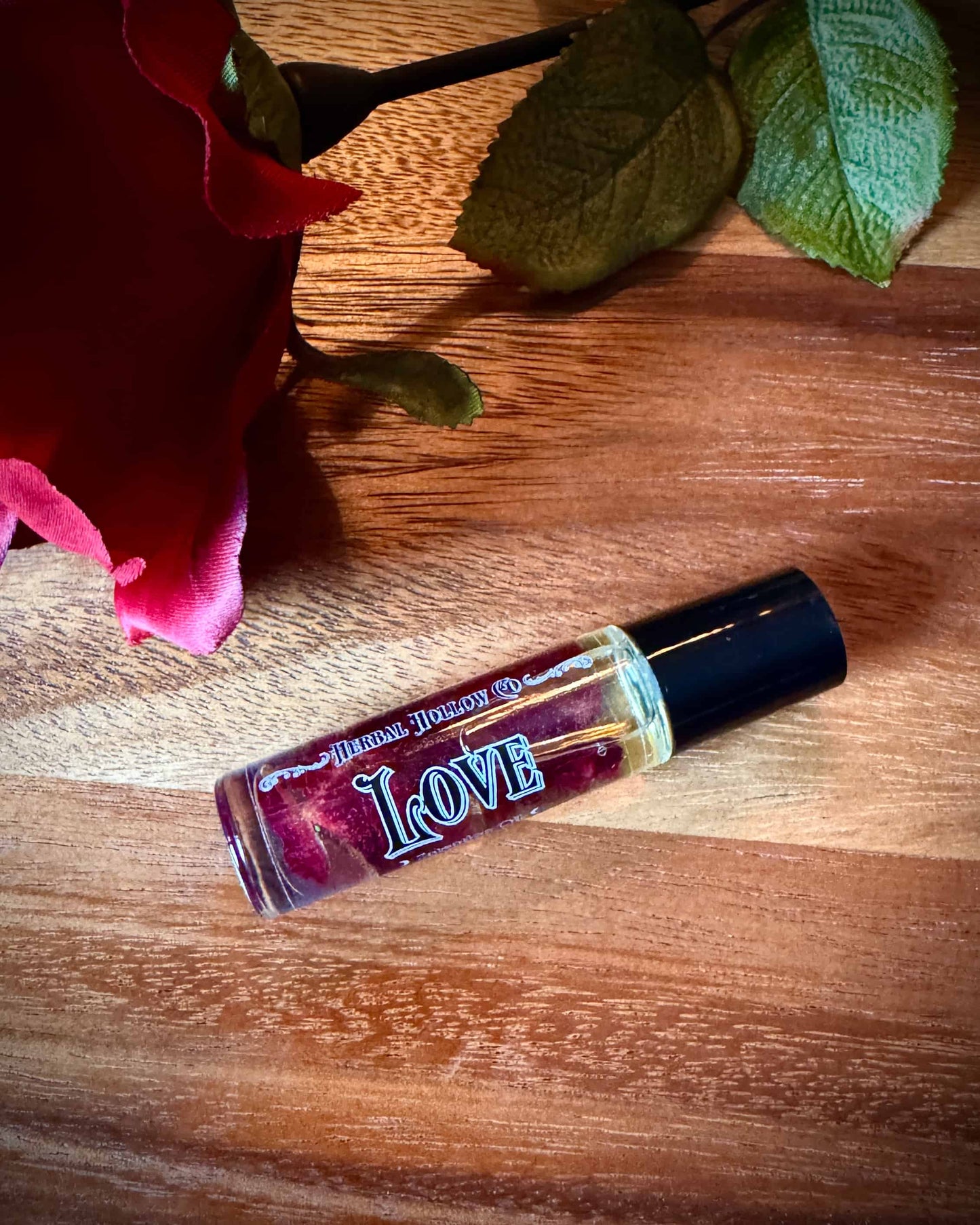 Small bottle labeled 'Love' with rose petals suspended in oil inside of it next to a red rose on a wooden surface