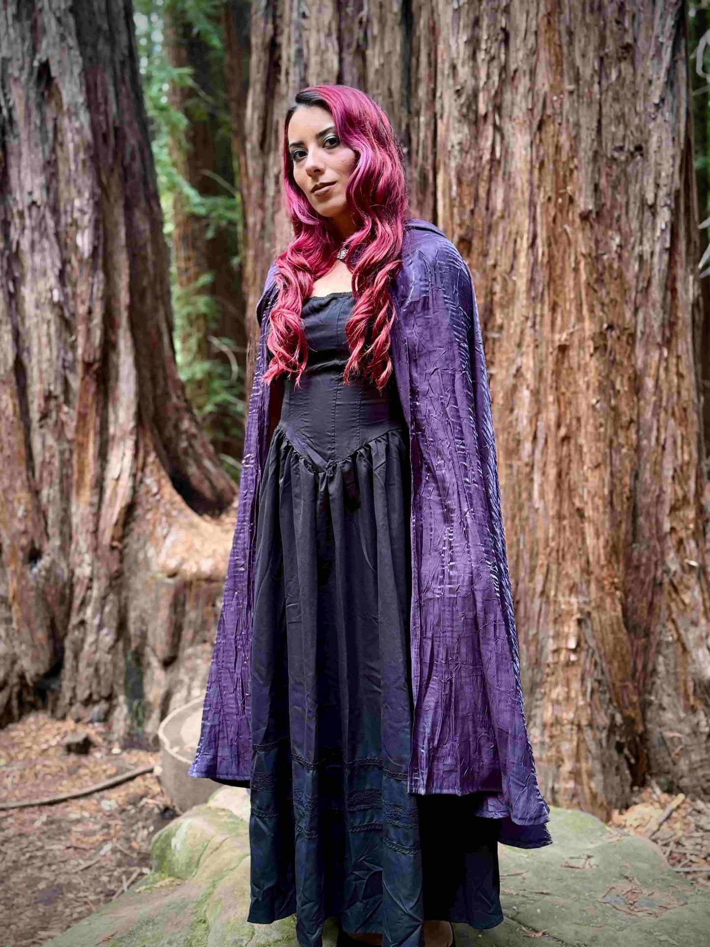 Woman with red hair wearing a long purple cape cloak and dark dress in a forest setting