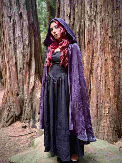 Woman with red hair wearing a purple cape cloak with a hood and a dark dress in a forest setting
