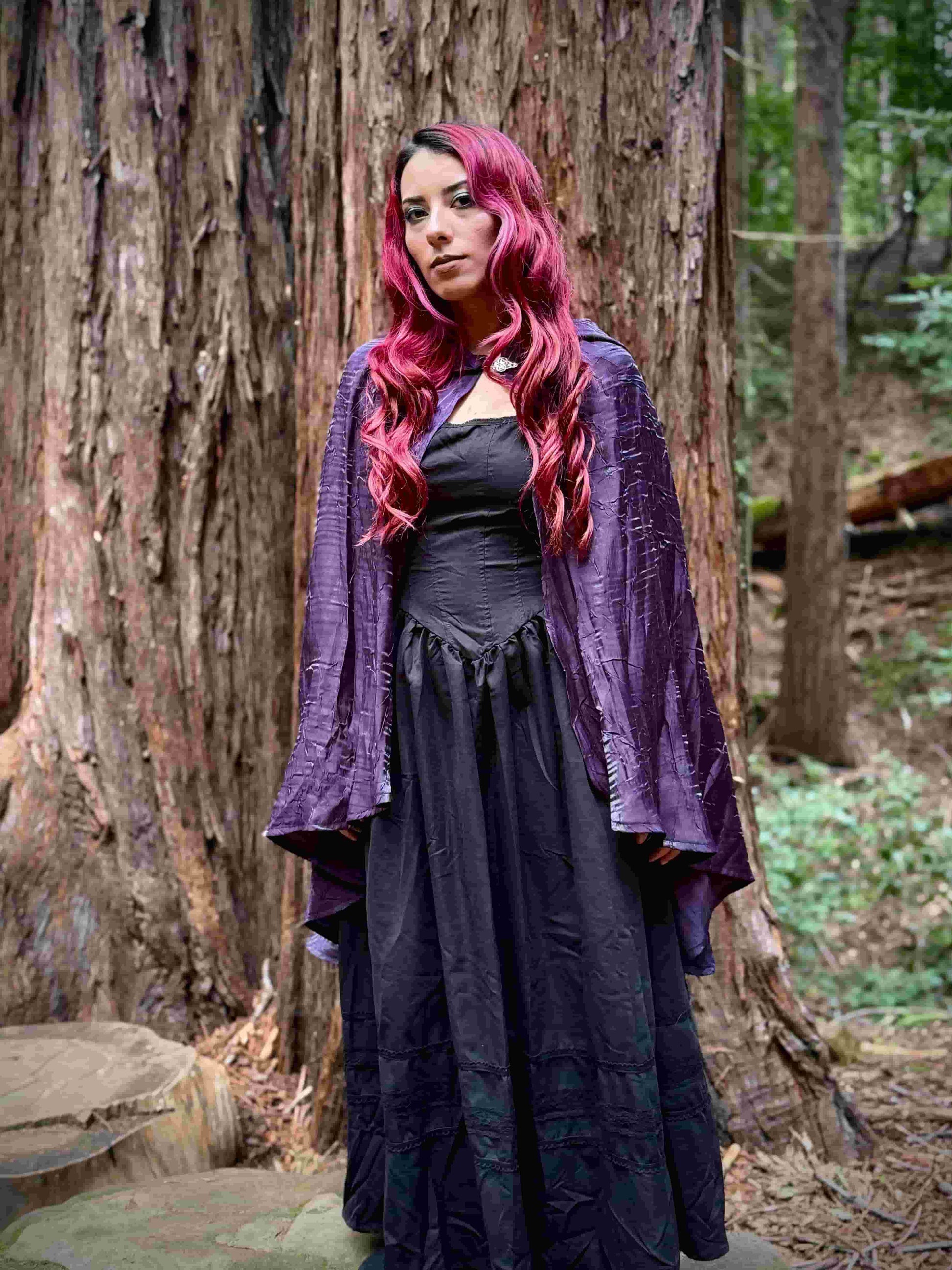 Woman with red hair wearing a purple cape cloak and dark dress in a forest setting