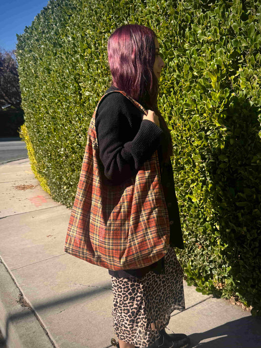 Person wearing a large plaid shoulder bag and leopard print skirt standing against a green hedge.