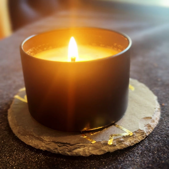 Burning candle being set on top of a coaster that features a gold image of the Herbal Hollow Co logo.