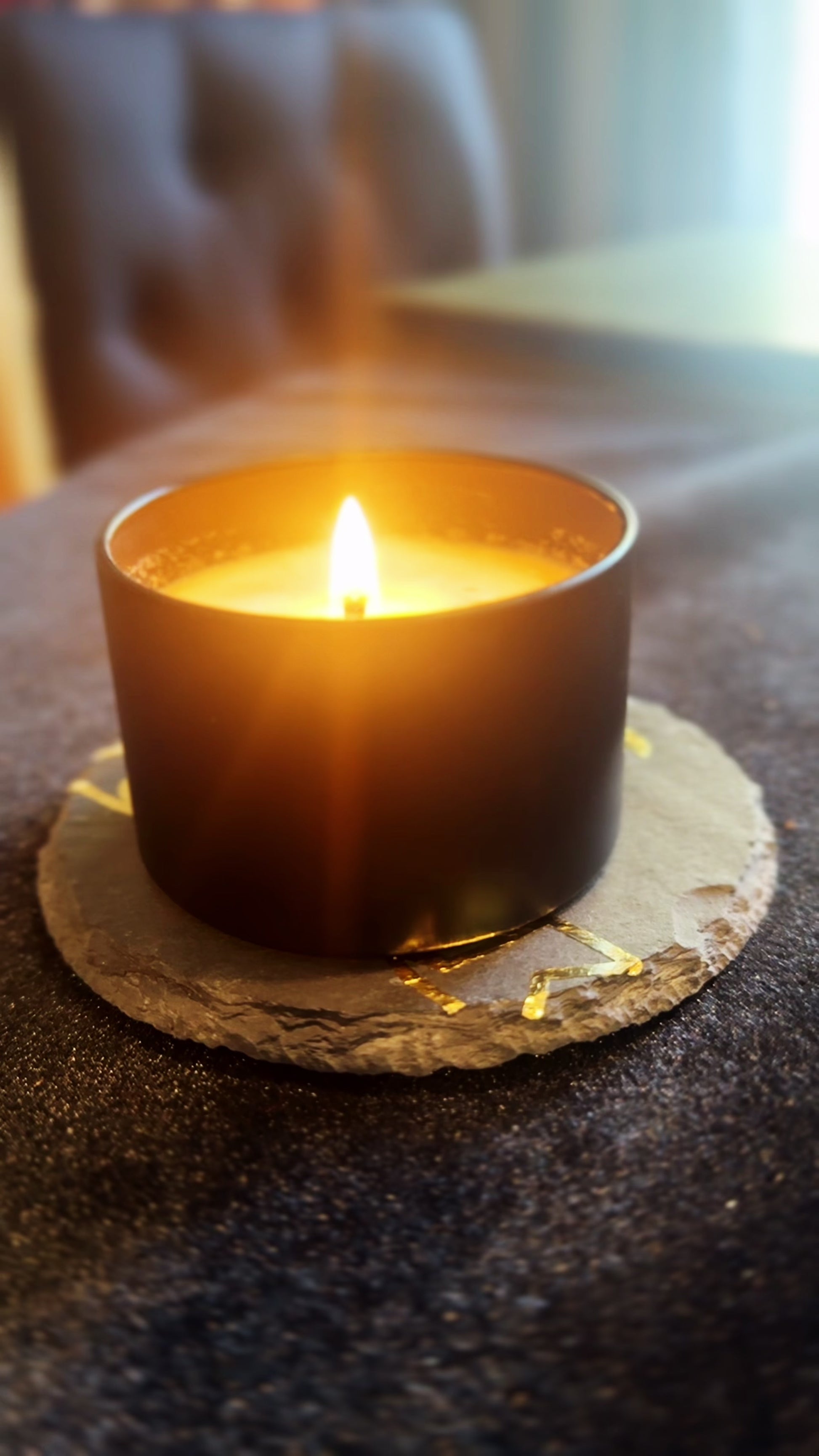 Burning candle being set on top of a coaster that features a gold image of the Herbal Hollow Co logo.