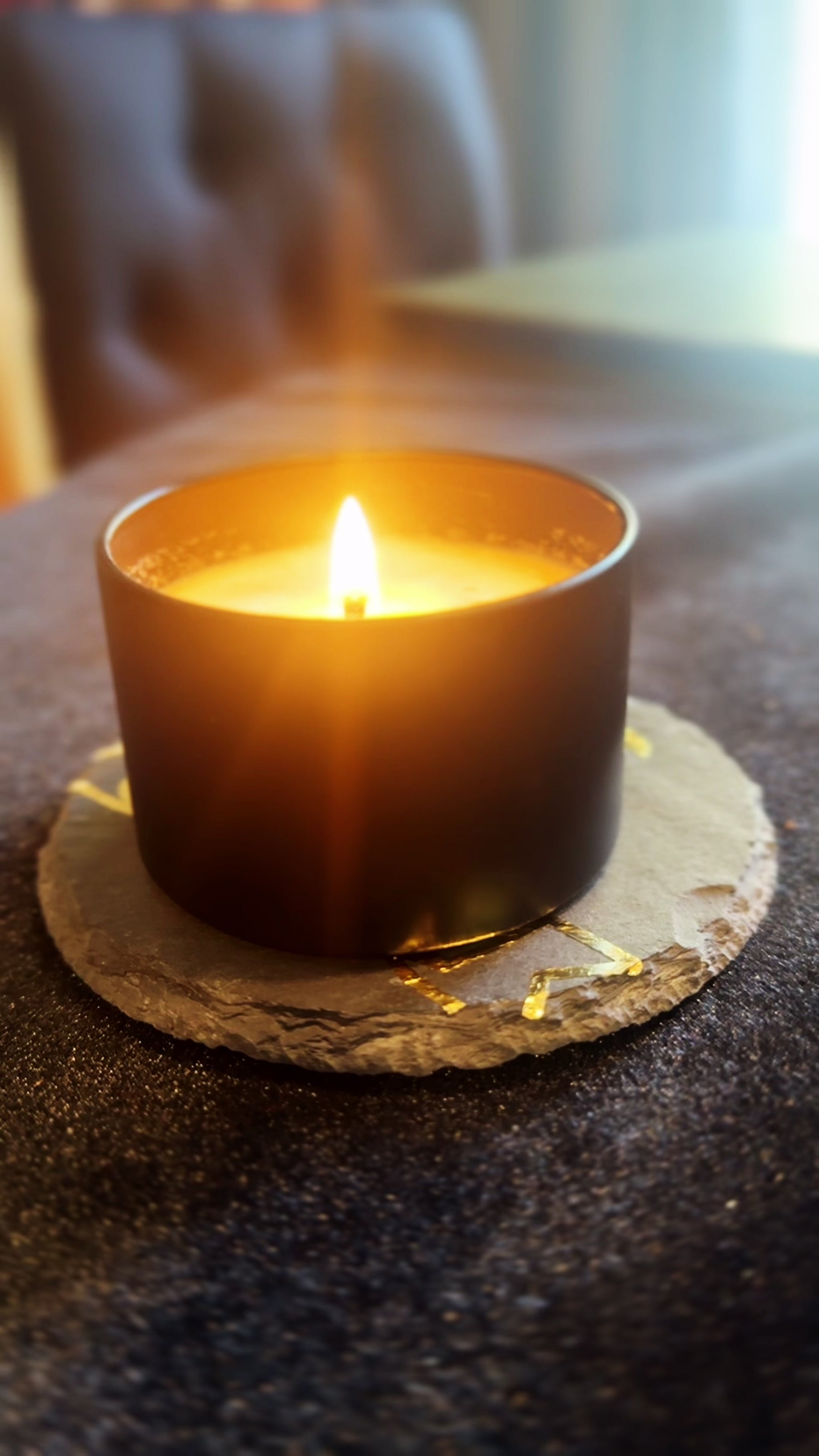 Burning candle being set on top of a coaster that features a gold image of the Herbal Hollow Co logo.