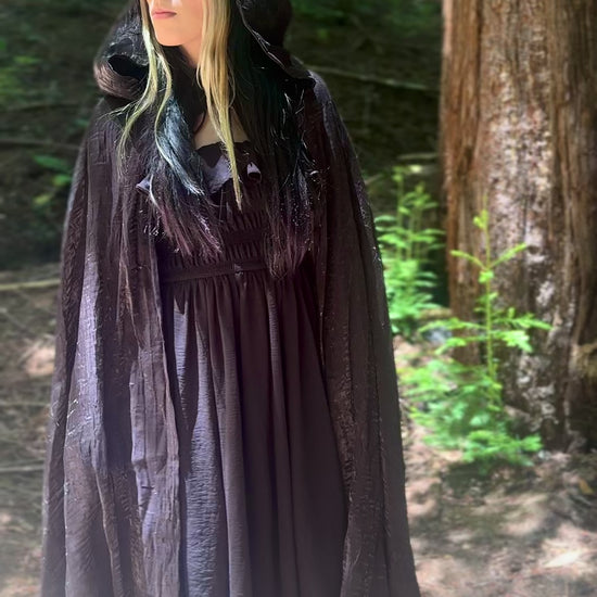 Person in a forest wearing a dark black hooded cape looking in the distance and walking