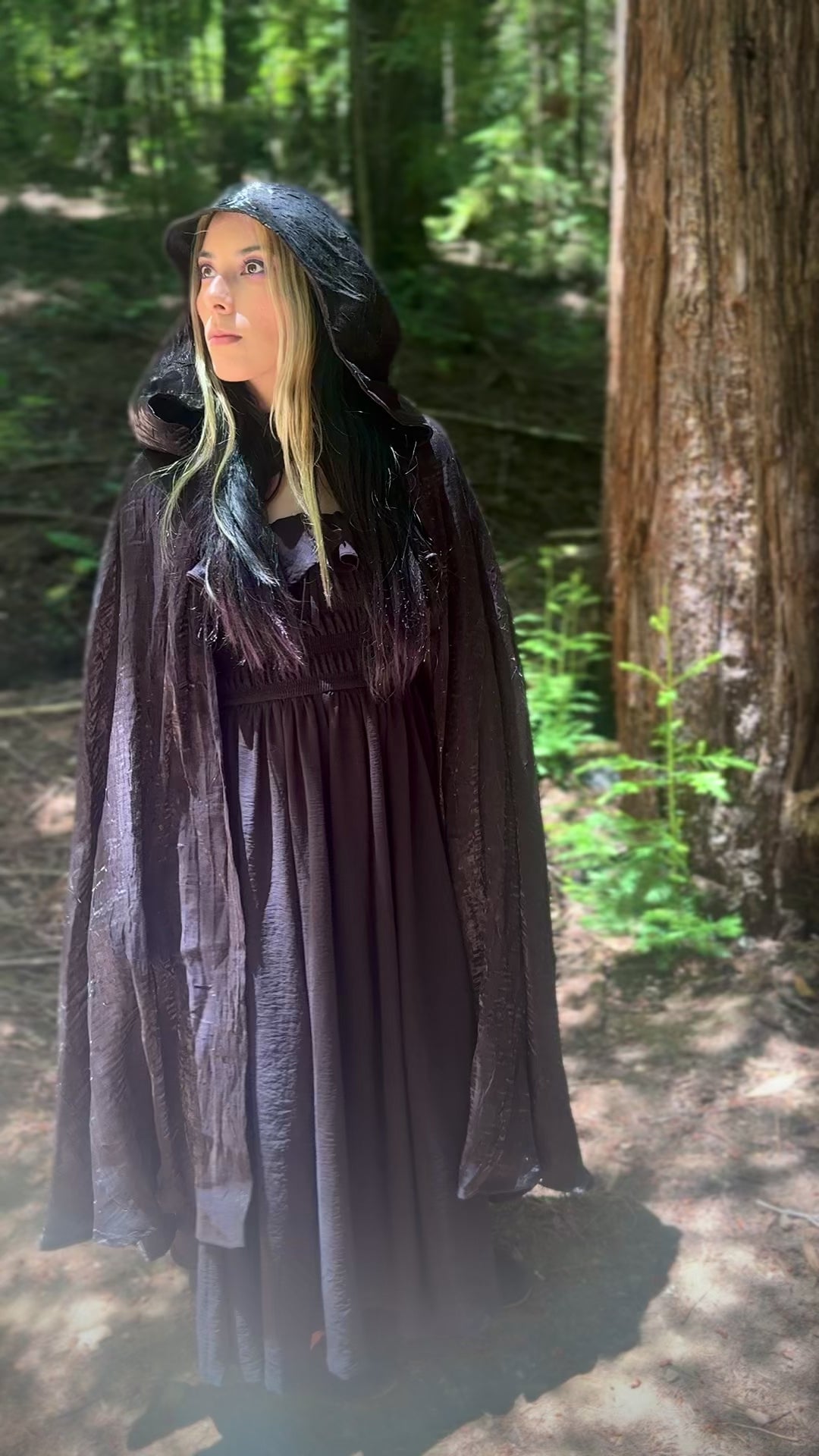 Person in a forest wearing a dark black hooded cape looking in the distance and walking